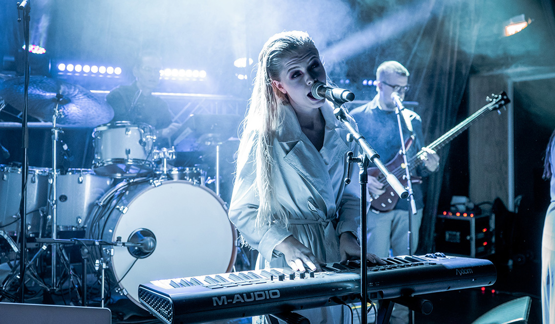 En jente står på en scene med lyseblått/hvitt lys i bakgrunnen. Hun har lagt blondt hår og har på seg en hvit kjole. Hun synger inn i mikrofonen og spiller keyboard.