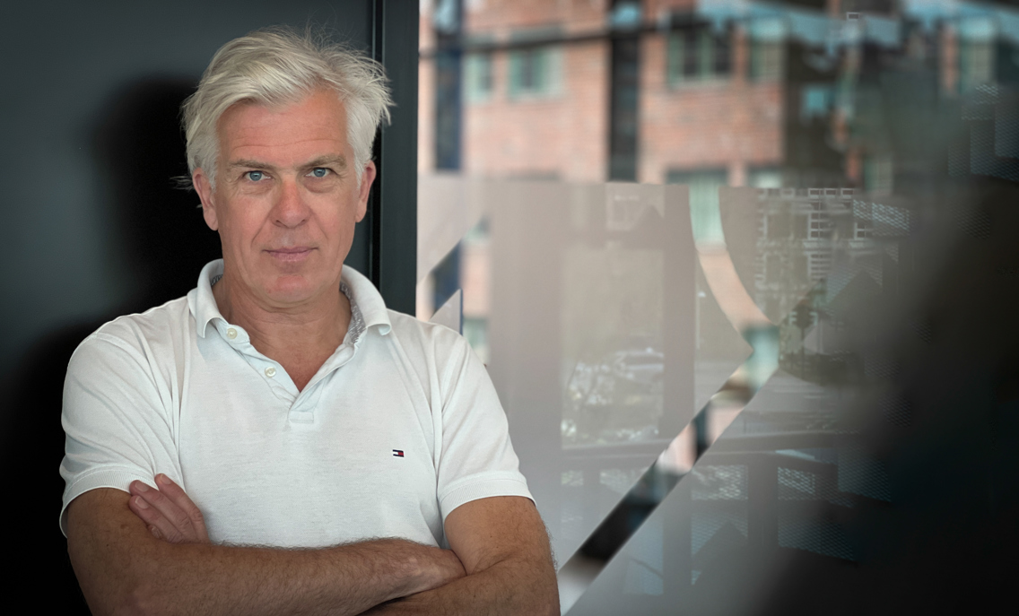Jørn Mortensen har på seg hvit skjorte og står foran en grå vegg og et vindu. Han ser inn i kamera med armene i kors.