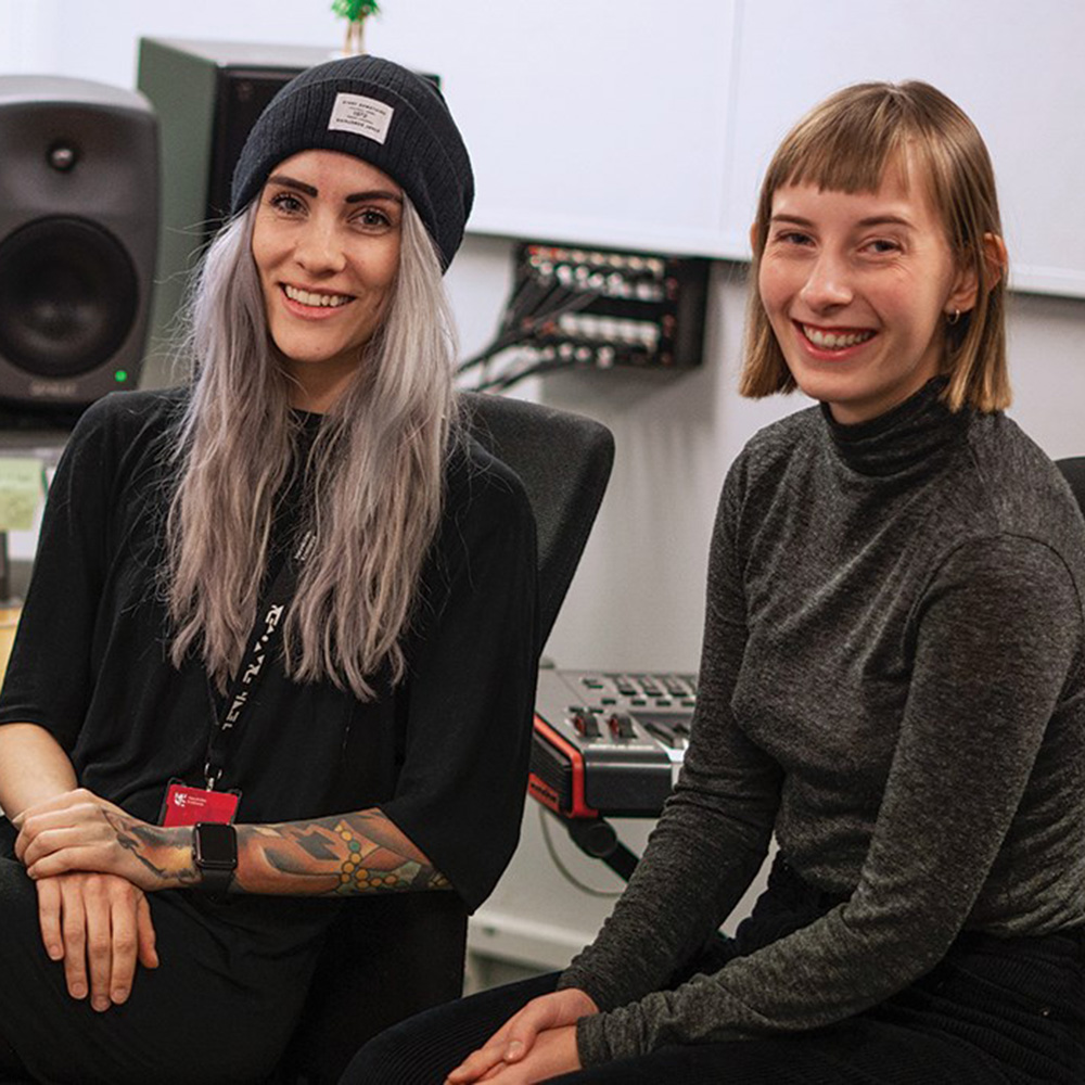 Ida Iki og Oda Tilset sitter i lydstudio på Fagskolen Kristiania. Ida har blondt langt hår, tatoveringer og lue. Oda har kort hår og pannelugg, og en grå høyhalset genser.