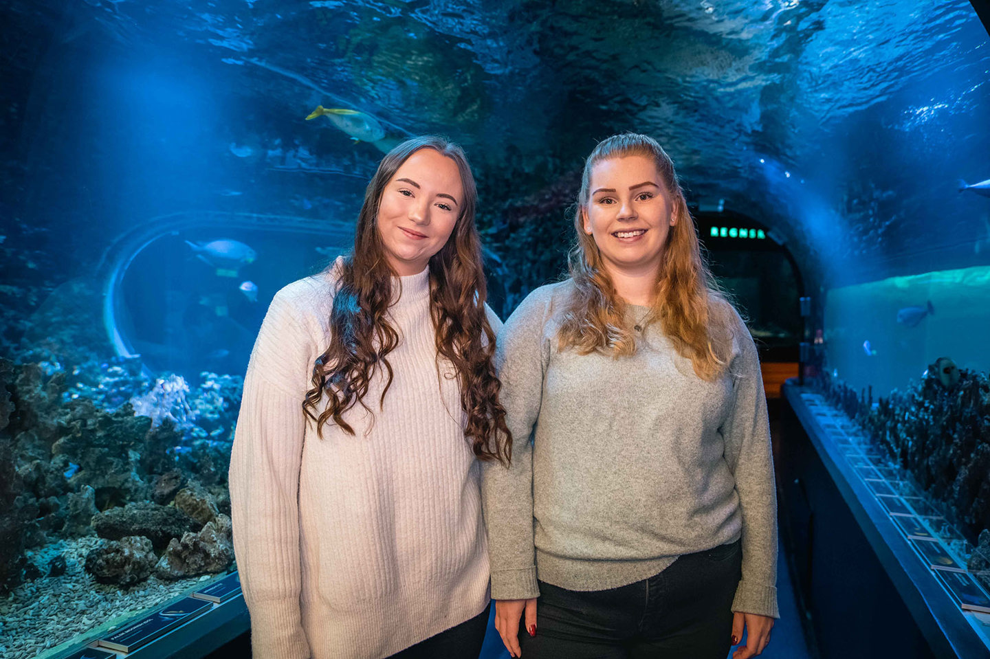 To jenter står og smiler i en vanntunnel med fisker rundt