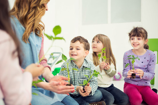 Tre barn smiler og holder små planter i et klasserom mens en voksen i en denimskjorte gir veiledning.