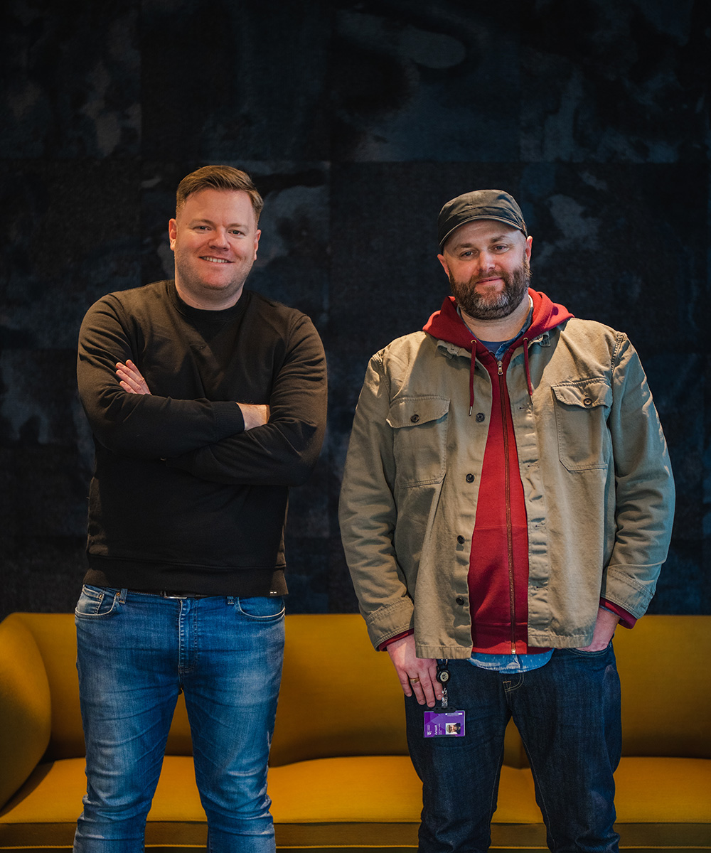 Foto av Chris Haaland og Scott Baker-Marflitt som står ved siden av hverandre og smiler til kamera. Bak dem står en gul sofa. 