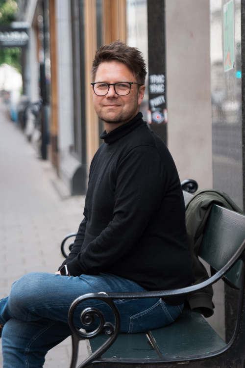 Odin sitter på en benk langs gaten, med butikkvinduer i bakgrunnen.