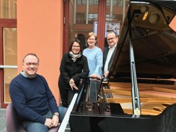 Foto av mann som sitter ved et piano, to kvinner og en mann i bakgrunnen.