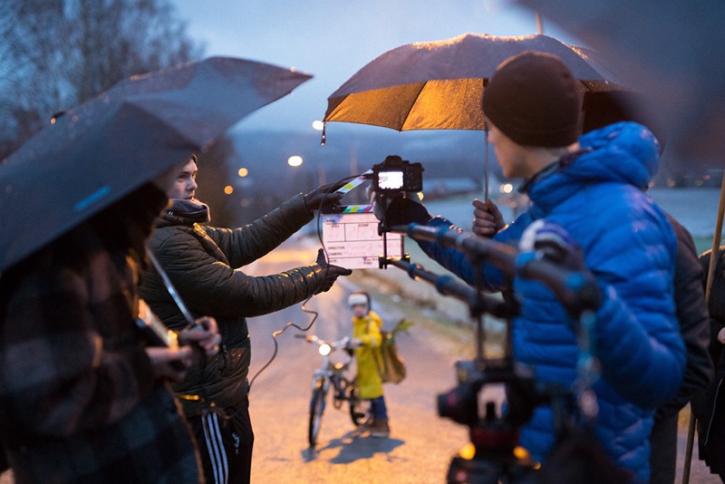 Bilde av et filmsett ute i regnvær. Flere på sett holder paraply over utstyr.