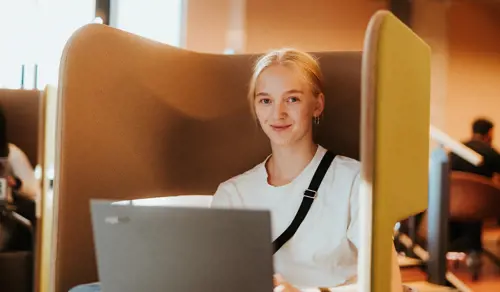 Jente sitter og jobber på en PC. Hun smiler lurt til kamera.