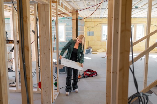 Marita bærer en plate i et bygg under oppføring, omgitt av reisverk og byggematerialer.