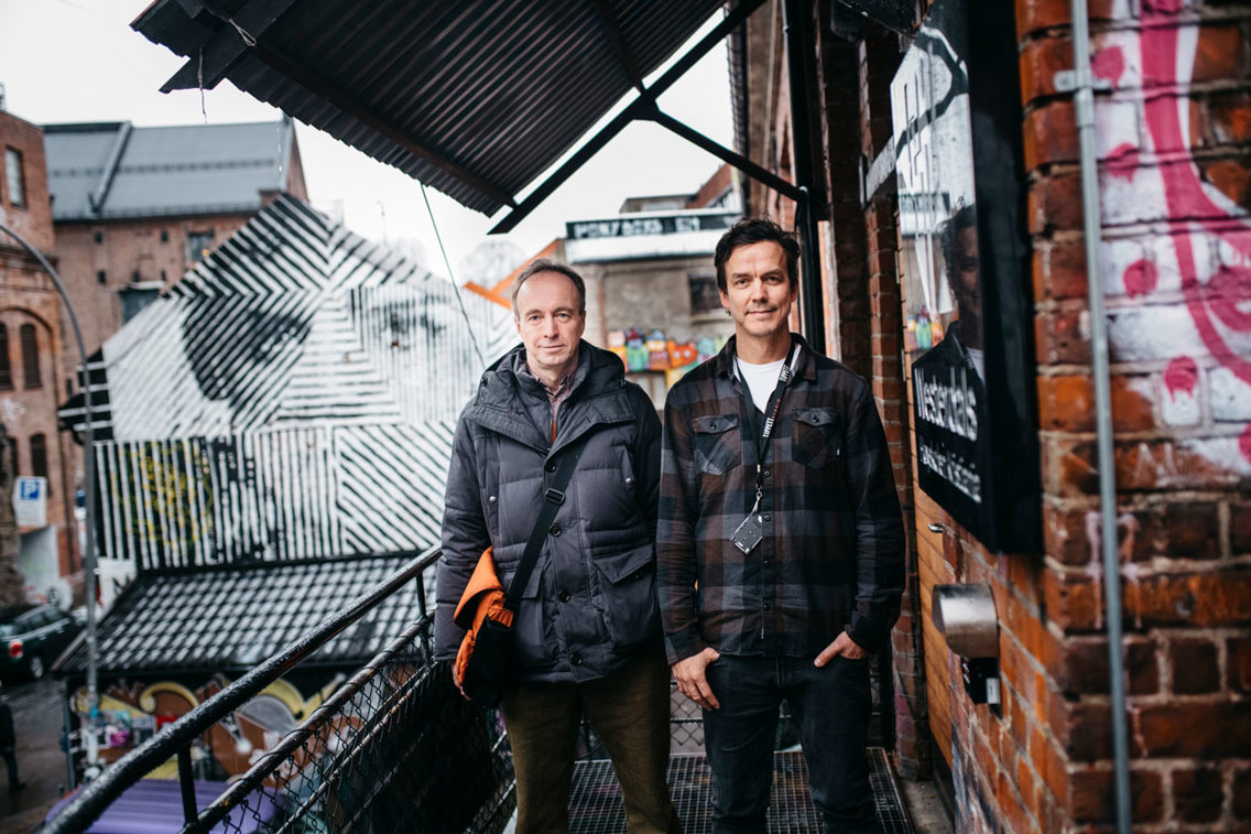 To menn står ved en gammel fabrikkbygning med grafitti.