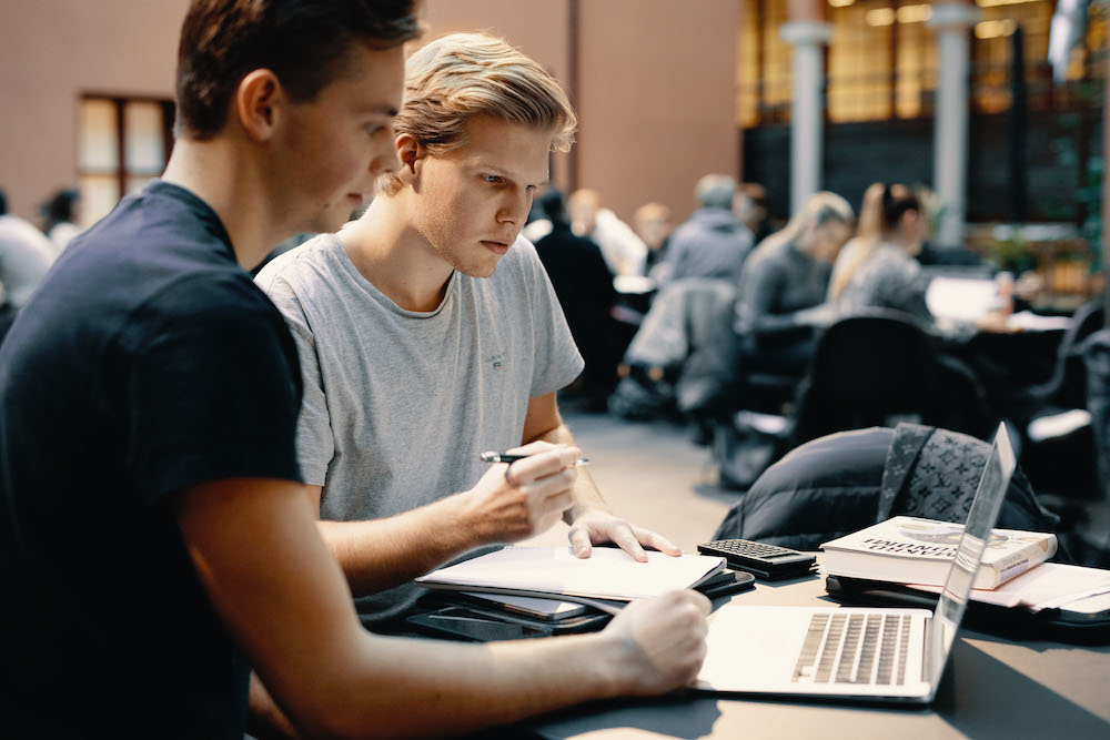 Foto av to mannlige studenter som ser på en laptop mens de skriver notater.