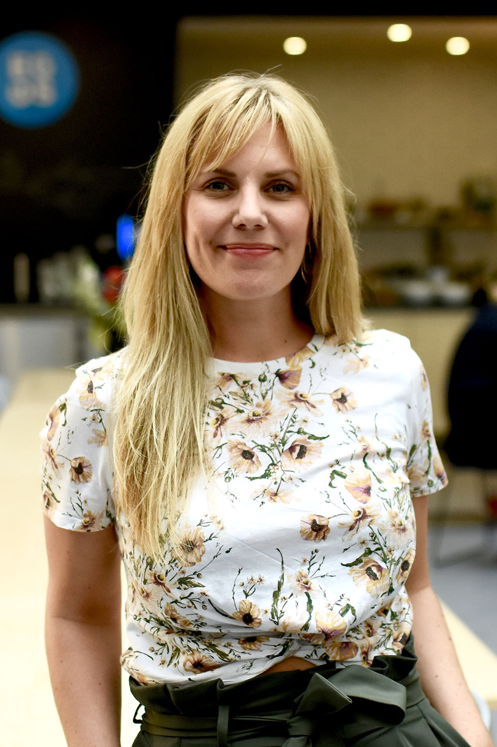 Trine Harnes har blondt, langt hår. Hun har på seg en hvit, blomstrete t-skjorte og en grønn bukse. Hun har hendene i lommen og smiler til kamera.
