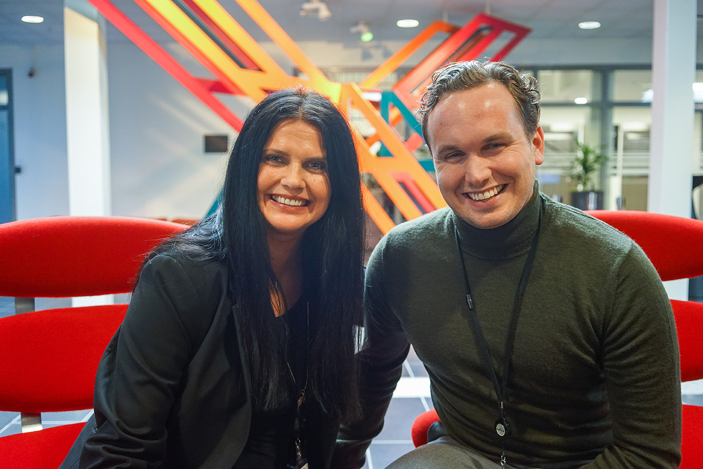 Foto av Anette Løland og Henrik som sitter sammen i hver sin stol. Anette har mørkt hår og Henrik lyst, begge smiler.