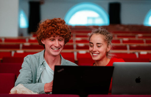 To unge personer, en med rødt krøllete hår og en med blondt krøllete hår, smiler og ser på en bærbar maskin sammen i et auditorium med røde seter i bakgrunnen.
