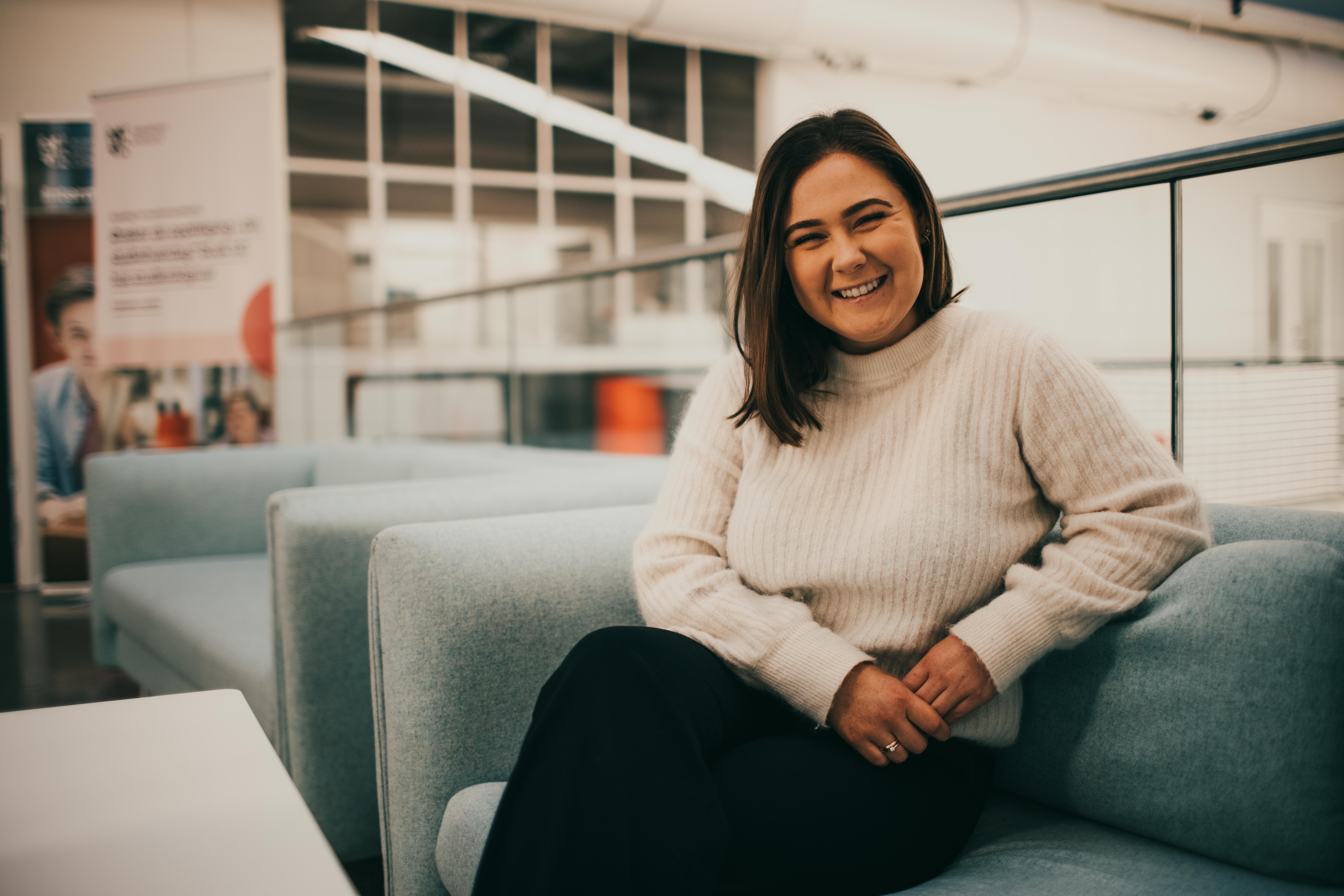 Masterstudent Lydia i hvit genser og sort bokse. Sitter i blå sofa på fjerdingen campus, Høyskolen Kristiania.