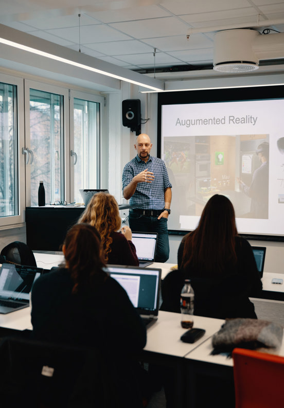 Bildet viser en mannlig foreleser som står foran en gruppe studenter i et klasserom. Han står ved en skjerm som viser ordene "Augmented Reality". Studentene, som hovedsakelig er sett bakfra, bruker bærbare datamaskiner, noe som antyder at de tar notater eller deltar aktivt i leksjonen. Klasserommet ser moderne ut, med lyse vinduer i bakgrunnen.