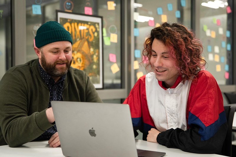 Foto av to menn som sitter rundt en MacBook og diskuterer. 