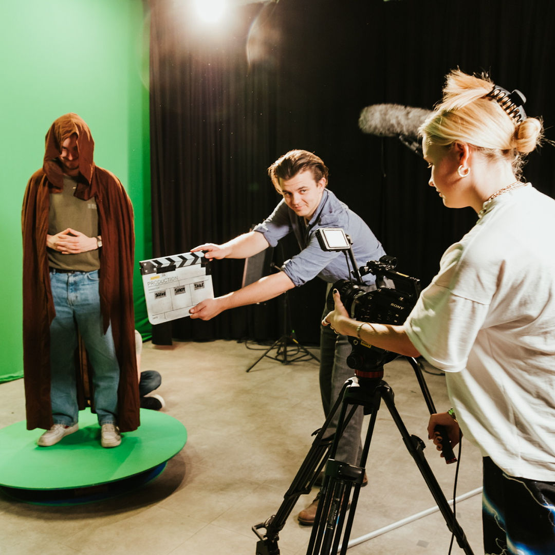 Vi er på et greenscreen rom på fagskolen. Tre studenter er med, ei som fører kamera og to gutter som står foran.
