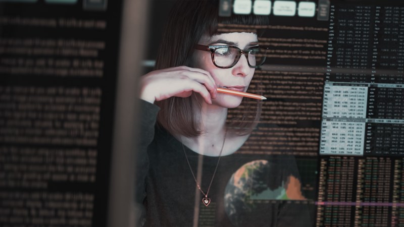 Kvinne med briller som sitter med en blyant ved munnen, og ser på en skjerm med koder og tall. 