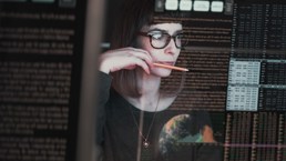 Kvinne med briller som sitter med en blyant ved munnen, og ser på en skjerm med koder og tall. 