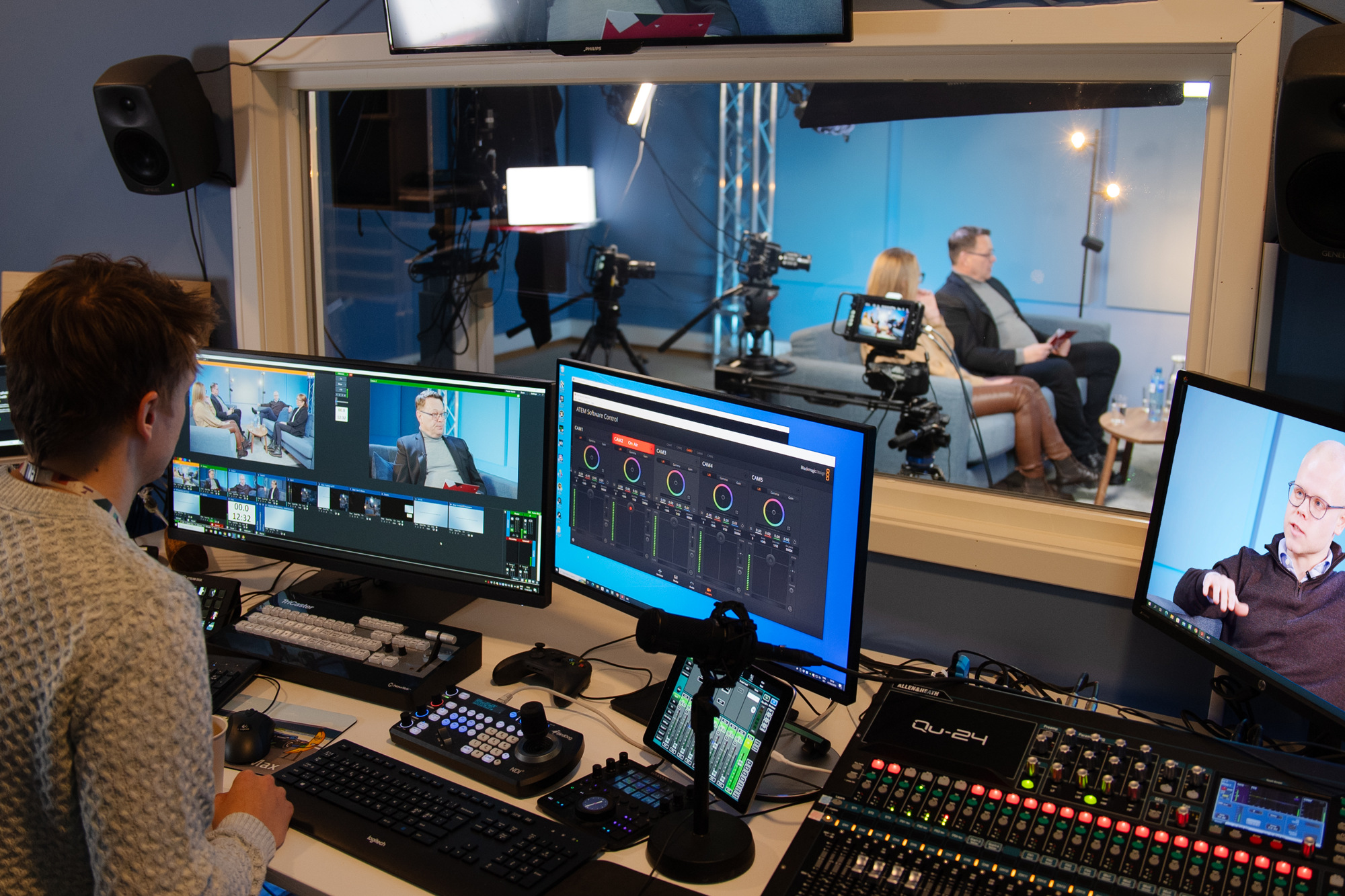 En person i studio ser på skjermer som viser to personer i sofa under opptak.