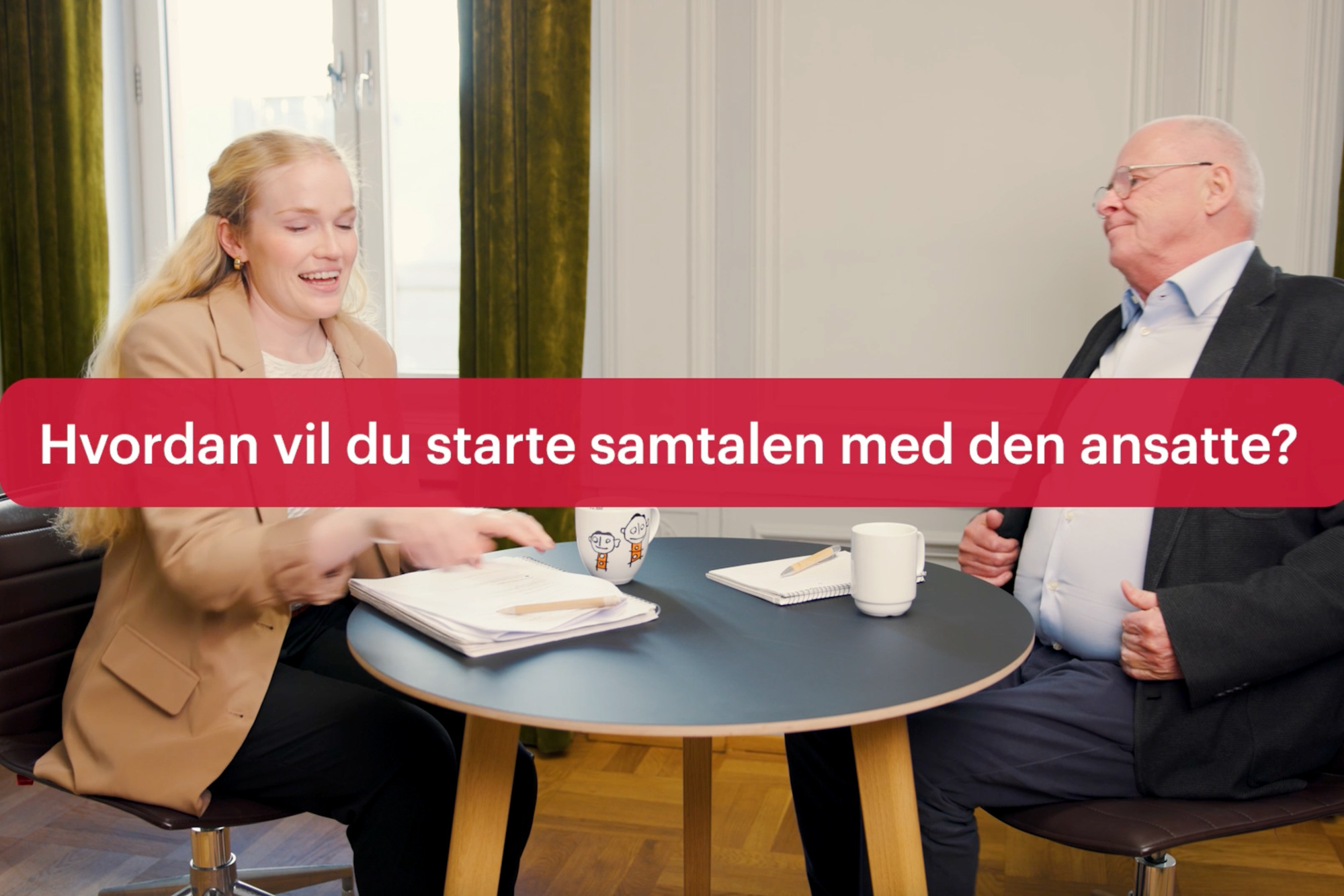 En mann og en kvinne sitter på hver sin side av et rundt bord inne på et møterom. 