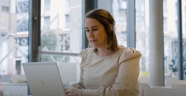 Miniatyrbilde fra videosnutt. En person med langt brunt hår, konsentrert om arbeid på en bærbar datamaskin i et lyst kontormiljø med store vinduer og en urban bakgrunn.
