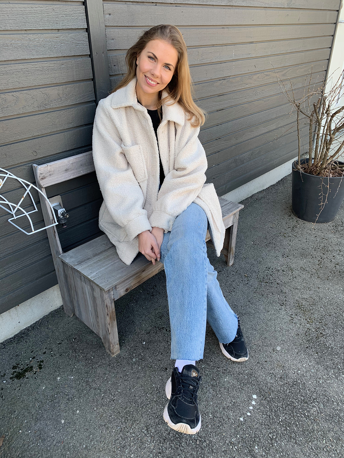 Camilla Lindström har blondt hår og har på seg en hvit jakke, svart genser, Adidas joggesko og blå jeans. Hun sitter på en liten benk og smiler.