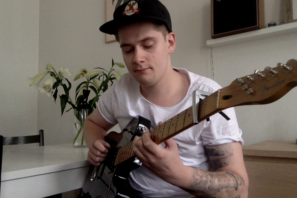 Olav er HK-student og sitter med gitaren, fokusert. Har på seg caps og hvit tshirt.