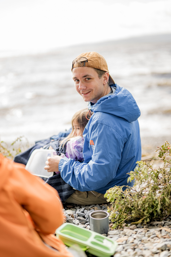 En person sitter ved vannkanten og spiser sammen med et barn ved sjøen