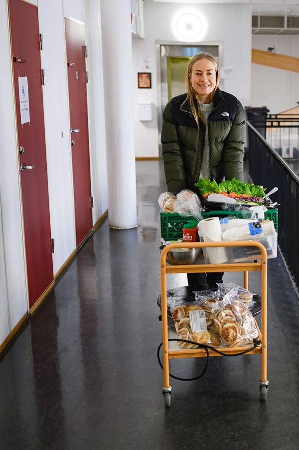 Foto av Susanne Tiller med handlevogn full av frukt og grønt, gående gjennom en gang i et skolebygg.