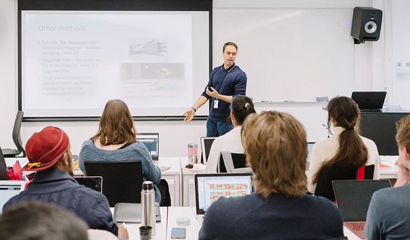 Foto av mannlig foreleser foran en tavle, som underviser en klasse.