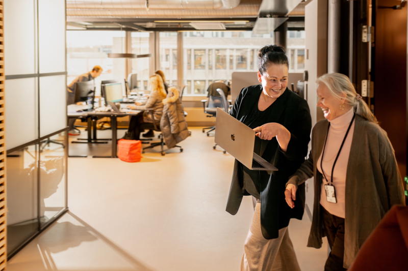 Personer går gjennom et åpent kontorlandskap med arbeidsplasser, én holder en bærbar PC og snakker med en kollega