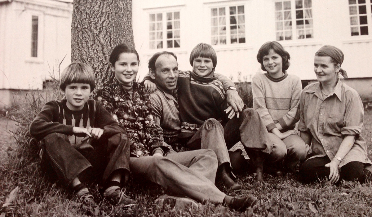 Bildet viser et gammelt fotografi i brune toner. En familie sitter på en gressplen under et tre foran et hvitt hus. 