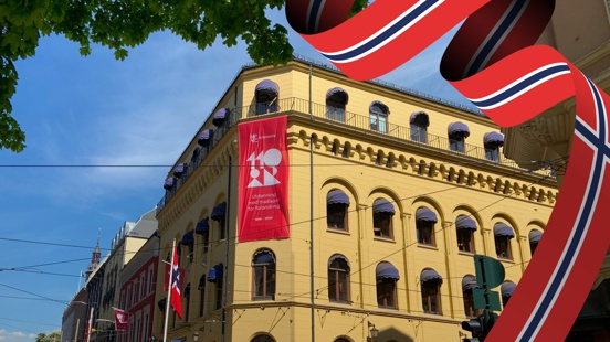 Bildet viser fasaden på hovedkontoret, med et banner som henger øverst med 110-års emblemet til Kristiania, mens bildet omkranses av norske flagg og grønne grener. 