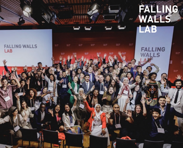 Deltagere på konkurransen i Berlin står samlet med hendene i været foran en vegg med logoen til Falling Walls.