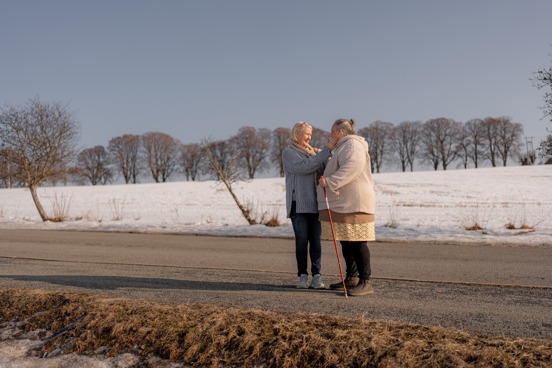 To personer er i en samtale og står på en vintervei, hvor den ene støtter seg med gåstav.