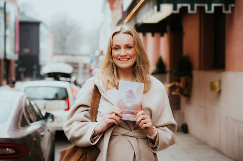 Ingvild står i en gate og smiler mens hun holder romanen hun har skrevet.