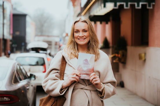 Ingvild står i en gate og smiler mens hun holder romanen hun har skrevet.