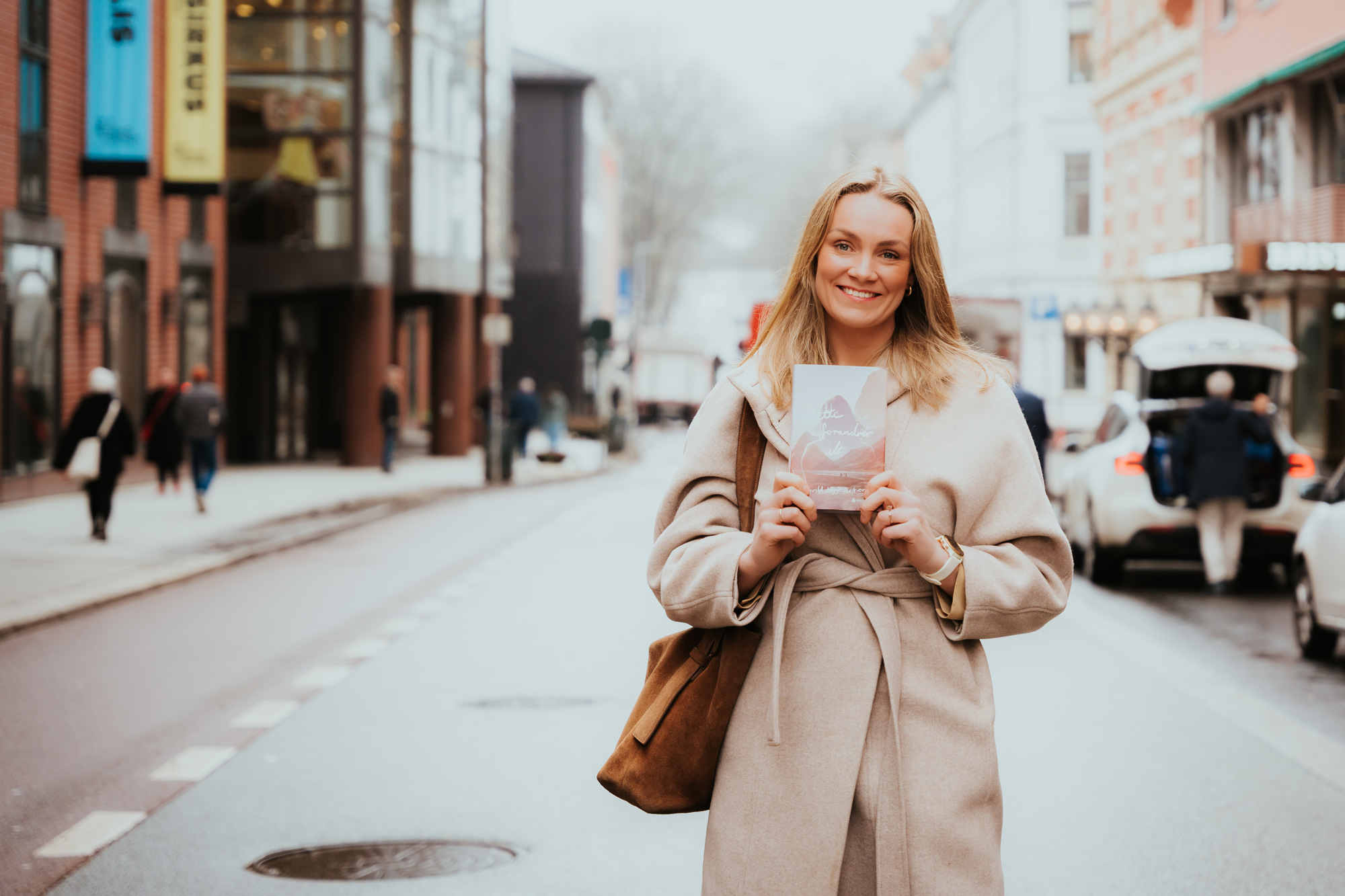 Ingvild står å smiler på gaten mens hun holder romanen hun har skrevet. 