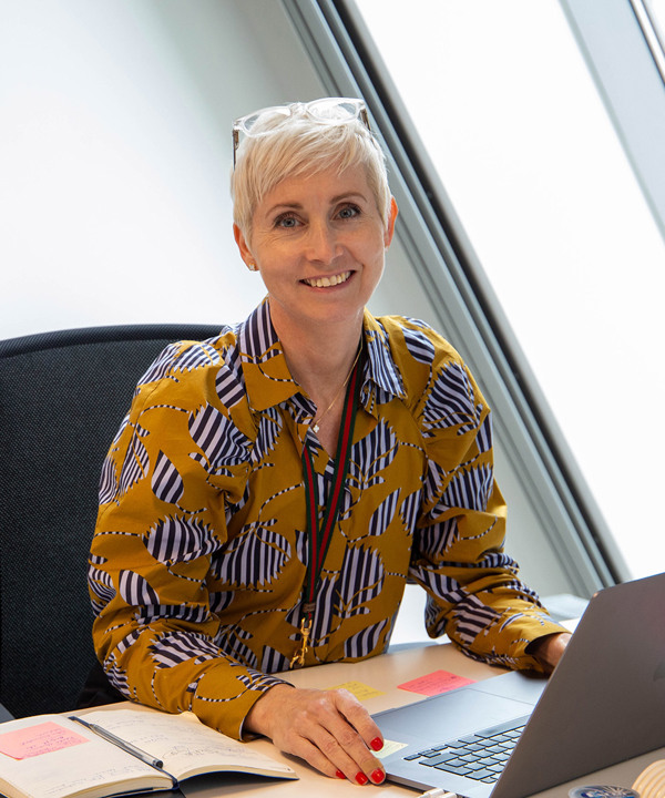 Avdelingsleder ved ESMOD Oslo, Elinor Johanne Hess, har kort blondt hår, transparente briller på hodet, sennepsgul skjorte og rød neglelakk. Hun sitter ved kontorpulten med en Mac fremfor seg.