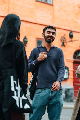 En liten gruppe studenter prater og ler. Til venstre i bildet ser vi en kvinne bakfra, hun har langt mørkt hår og Kristiania-handlenett over sin høyre skulder. I midten av bildet ser vi en mann med gråblå ribbestrikket genser med v-hals. Han har kort hår og skjegg. Han har også et Kristiania-handlenett over sin høyre skulder. 