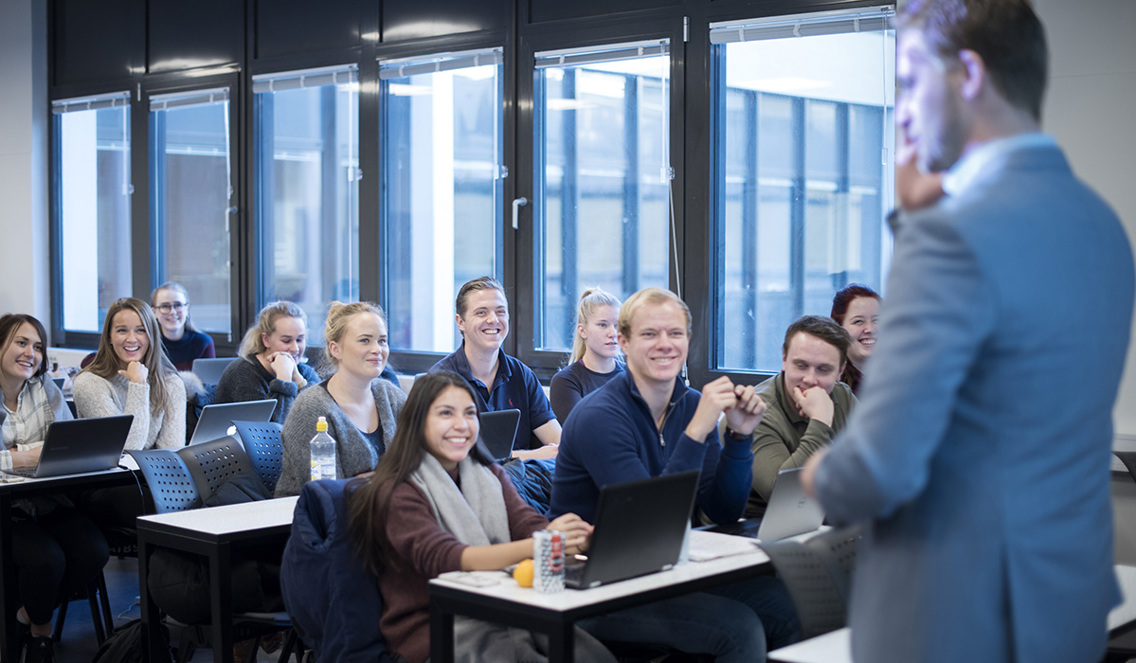 Bildet viser et forelesningsrom på Høyskolen Kristiania i Bergen. Studentene studerer Bachelor i markedsføring og salgsledelse. Studentene ser oppstilt opp på foreleseren som står helt i høyre i bildet. 