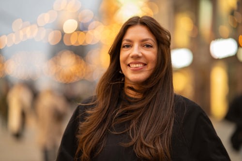 Kvinne med langt, tykt brunt hår smiler og ser inn i kameraet. Hun står i Karl Johans Gate en mørk vintermorgen og er omringet av koselig belysning