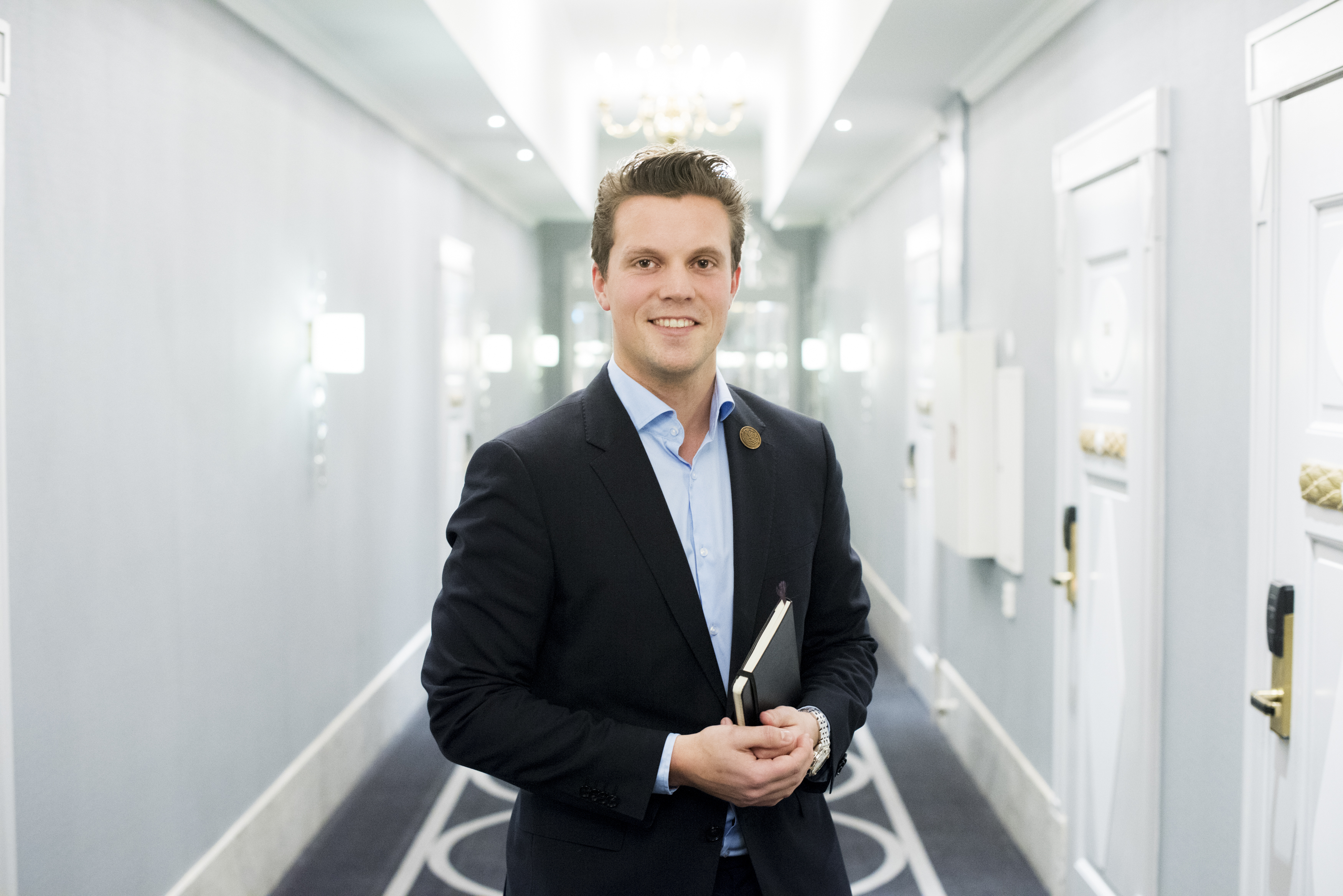 Foto av ung mann i dress som står i en hotellkorridor.