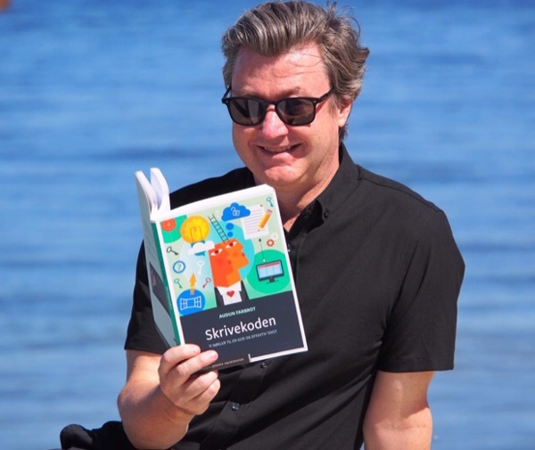 Mann med solbriller leser bok med havet i bakgrunnen.