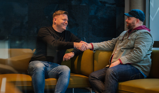 Foto av Chris Haaland og Scott Baker-Marflitt som sitter i en sofa og tar hverandre i hånden.
