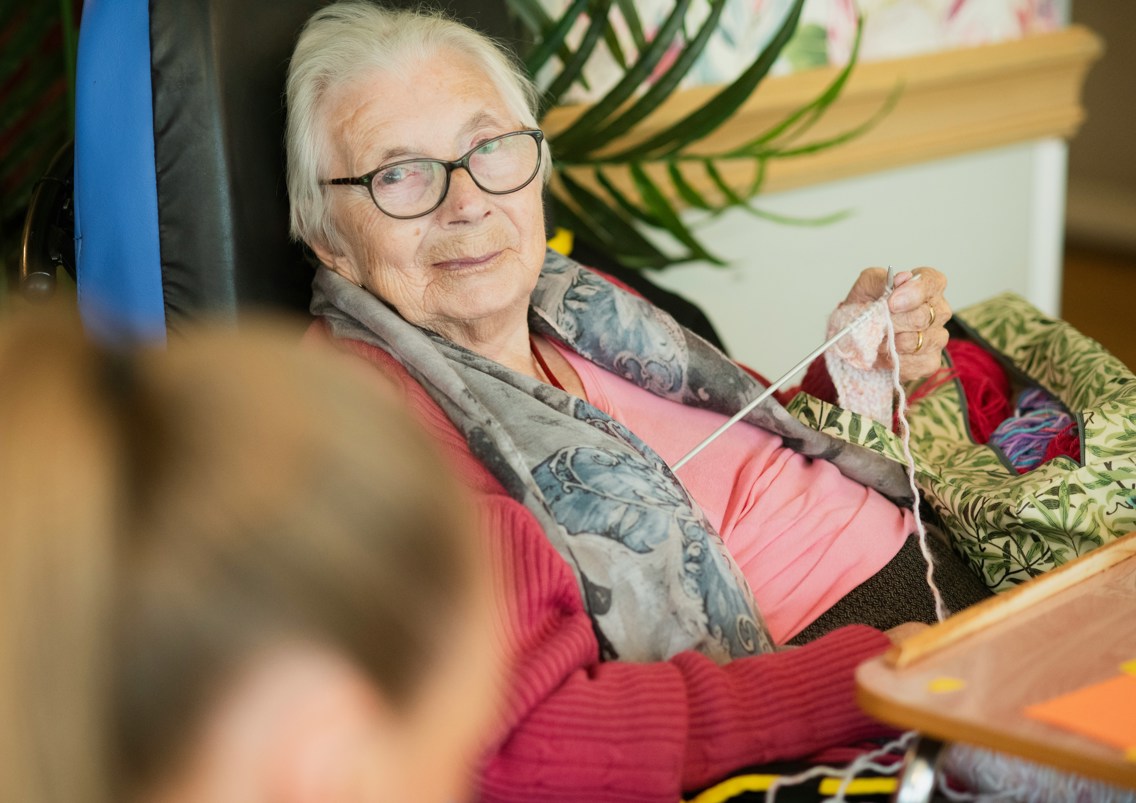 Eldre kvinne sitter og strikker og ser i kamera med en yngre kvinne ved siden av seg