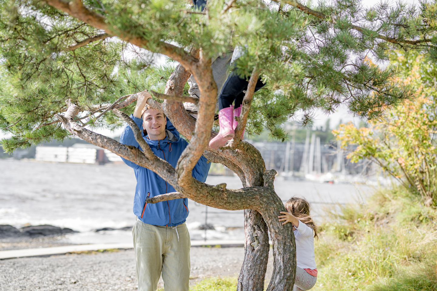 En voksen og to barn leker i et tre nær vannkanten.