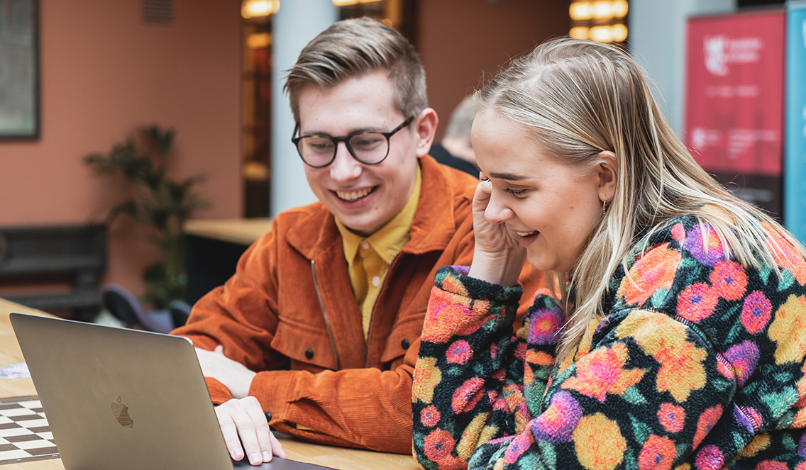 Joakim og Maren sitter på et høyt bord og ser ned i en Macbook med grått utseende. Joakim har sorte briller og har på seg en sennepsgul t-skjorte med en oransj jakke over. Maren har blondt hår og en blomstrete genser på seg. Hun hviler hodet i den ene hånden. Begge smiler.