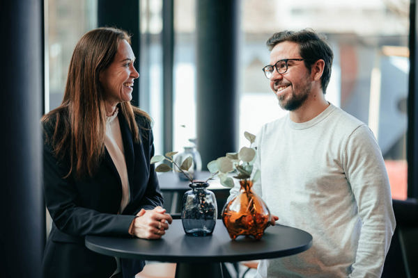 To personer, en kvinne og en mann, ler og ser på hverandre mens de står ved et bord med to vaser, i et rom med store vinduer.