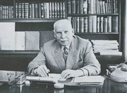 Foto i sort-hvitt av en mann som sitter ved en pult i et arbeidsrom med mange bøker i hylle.